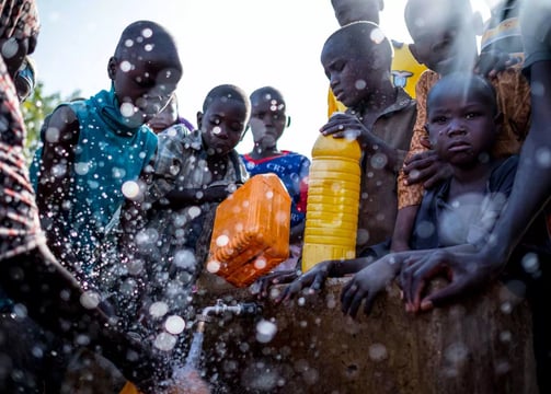 A group of African children and adults gathered around a water source, learning about hygiene