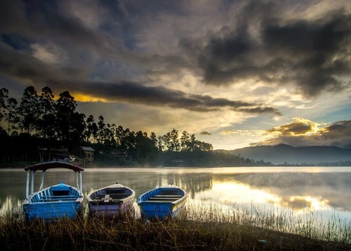 Tiga perahu bersandar di tepi Danau Situ Cileunca saat matahari terbit, dengan kabut tipis dan pantulan langit di permukaan a