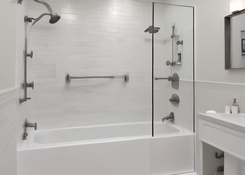 A modern bathroom features large, smooth tiles on the walls and floors, a sleek white countertop with built-in sink and storage cabinets, and a large illuminated mirror. Recessed ceiling lights provide ample illumination, and a walk-in shower with a glass partition is situated at the far end. A window allows natural light to enter, enhancing the minimalist aesthetic.