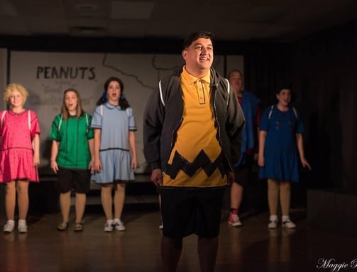 an actor playing Charlie Brown in a yellow sweater and black shorts standing in front of a group of