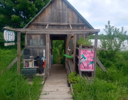 A wooden farmstand with an open entrance, featuring signs for organic eggs, free produce, and 'The D