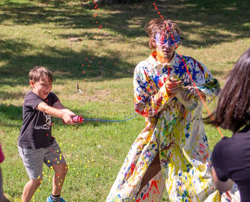 Summer Camp children shooting paint at the teacher in a white trenchcoat.