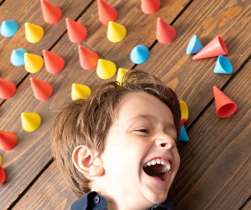 Laughing boy laying on the brown ground surrounded by blue, red and yellow silicone cones