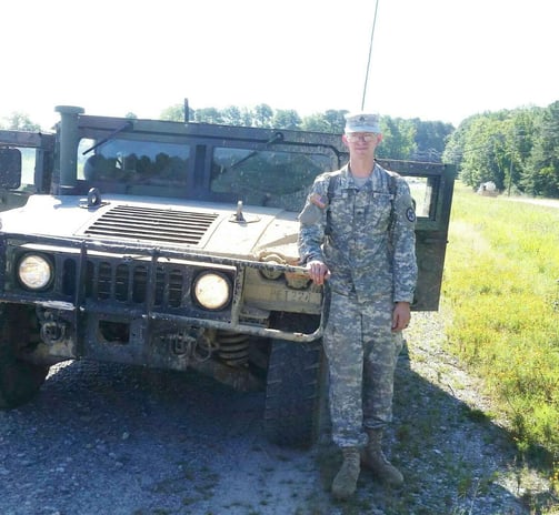 Tomkins in army uniform standing next to a green humvee covered in mud.