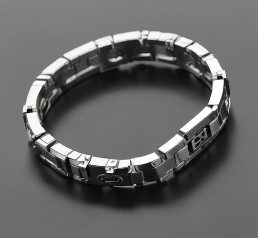 A close-up of a luxury watch with a silver and gold bracelet, featuring a gold bezel and a date display. The watch rests on a computer keyboard in an office setting, with a blurred background showing sunglasses, headphones, and a decorative pyramid.