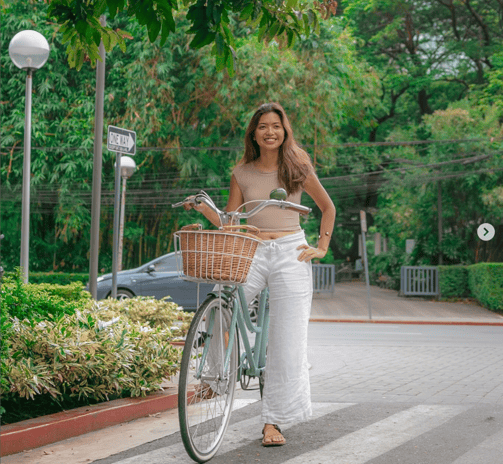 Femme souriante marchant avec un vélo de ville vert équipé d’un panier en osier, sur un passage piét