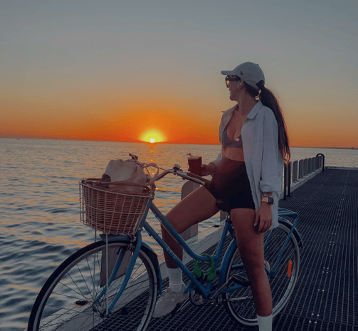 Femme posée sur un vélo bleu au bord de l’eau au coucher du soleil, boisson à la main.
