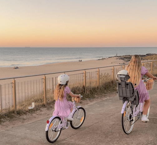 Mère et fille à vélo en bord de mer au coucher du soleil, toutes deux en robe violette.