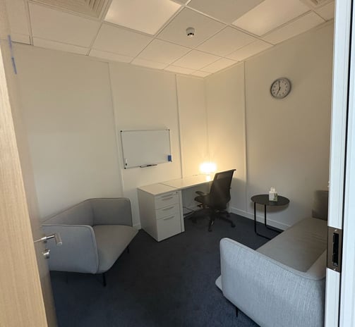 A therapy room, a grey armchair to the left with a sofa to the right, a desk on the back wall