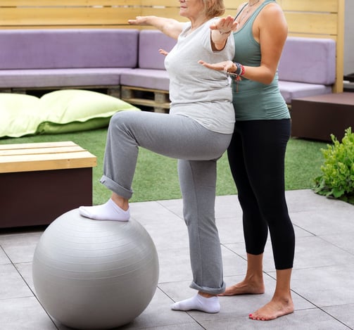 an elderly woman working on her balance using a yoga ball with the help of a trainer coach