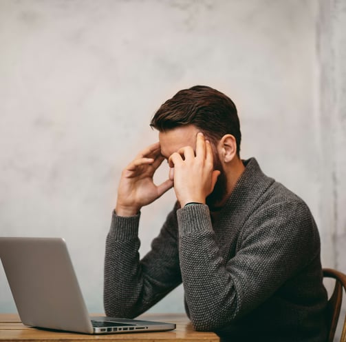 a man sitting at a desk with a laptop computer. holding his head with his hands