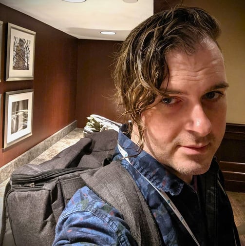 Photo of GJ, fair-haired adult with wavy, asymetrical hair and stubble, with backpack and lanyard in a conference center