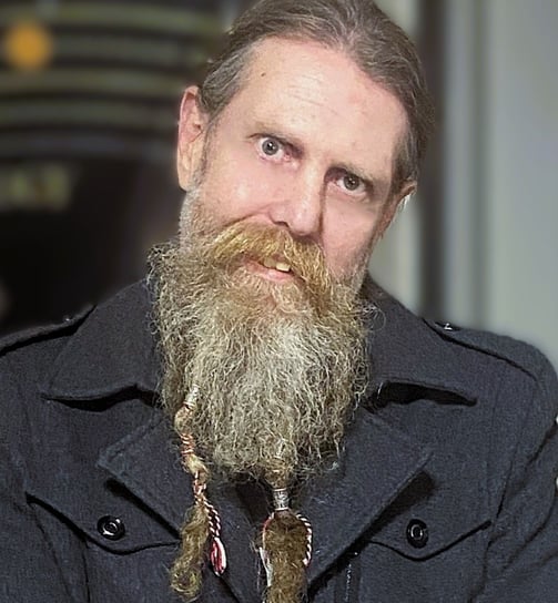 A man with a long beard is tilting their head to one side while wearing a well appointed coat