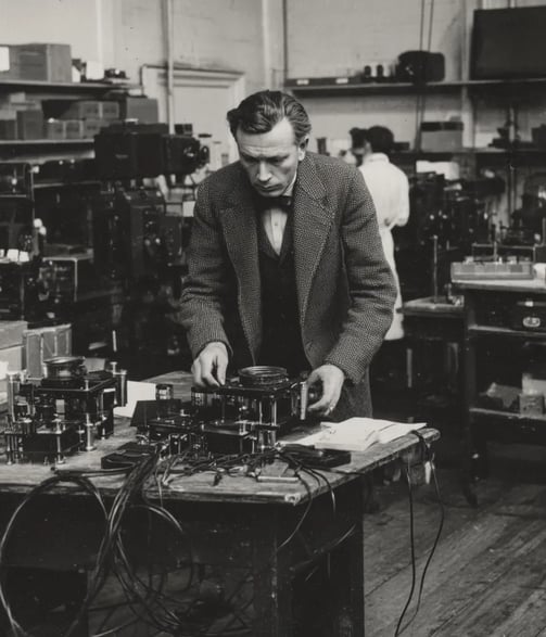 John Logie Baird in a tweed suit and bow tie adjusts a rotating Nipkow disc assembly
