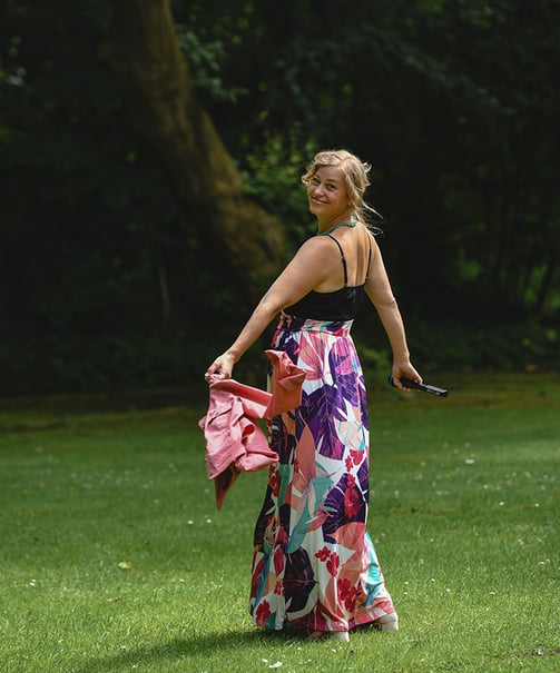  Das Bild zeigt eine lächelnde Frau in einem farbenfrohen, bodenlangen Rock mit floralen Mustern, der Pink-, Lila-, Türkis- u