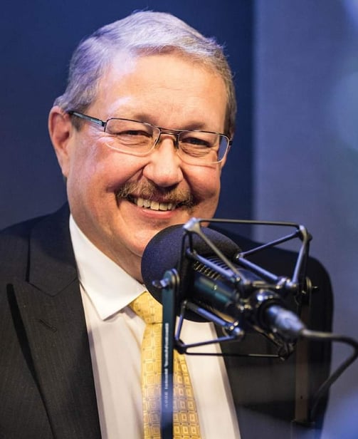 Man in suit and gold tie smiling behind a microphone.