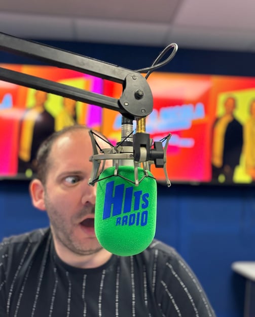 Alex Duffy is in a radio studio, face half-obscured behind a Hits Radio microphone.