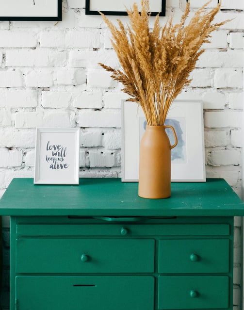 a green dresser with a vase of dried grass in front of a brick wall