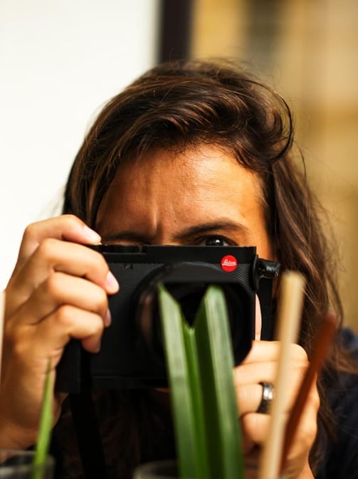 Close‑up of a person with a Leica camera, blurred green leaves in foreground - By ACAT Photos
