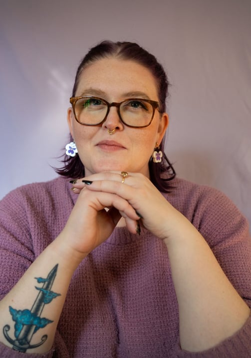 A photo of Kat in a purple sweater with pansy earrings and her hands steepled in front of her.