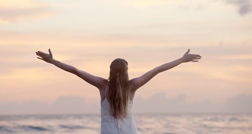 Lady facing her back and with hands lifted in the air. She faces the sea. 