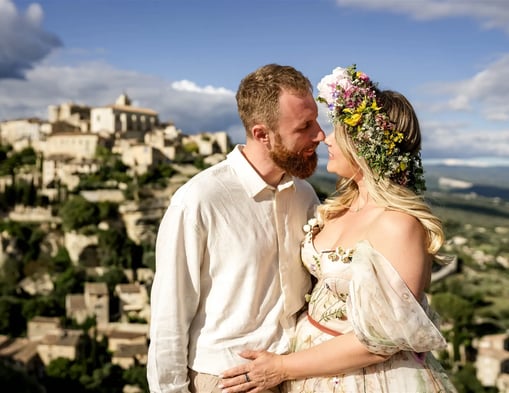Fine art couple session — bride with flower crown gazes at partner with Provençal village backdrop