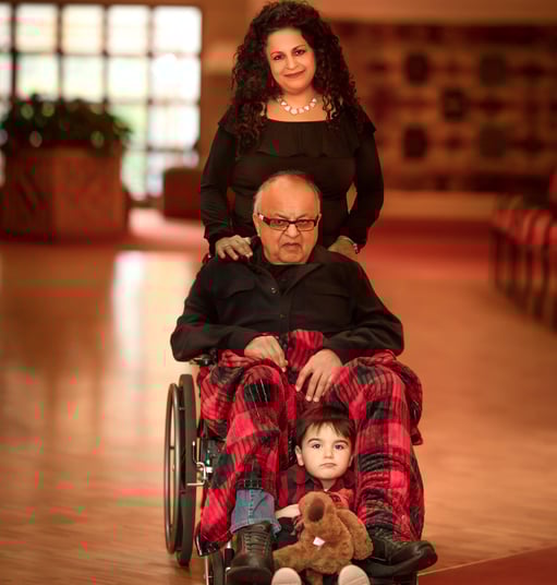 A woman pushes a frail man in a wheelchair, a young child holds a bear at the man's feet.
