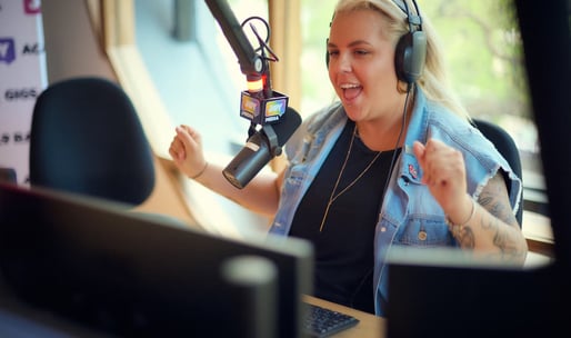 A blonde haired person with headphones on smiling into a microphone in a recording studio