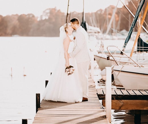 Hochzeitspaar steht auf einem Steg am Wasser, umgeben von Booten und Natur.