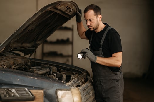 Mecânico levanta o capô de carro desgastado e inspeciona o motor com lanterna em oficina simples.