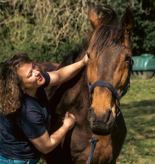 Gemma and a bay horse smiling in the sunshine whilst the horse is getting a massage on their neck.
