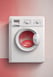 Two white front-loading laundry machines are placed side by side beneath a wooden countertop. The left machine appears to be a washing machine while the right one is a dryer. They are situated in a utility room with a grey floor and some plumbing fixtures visible to the right.