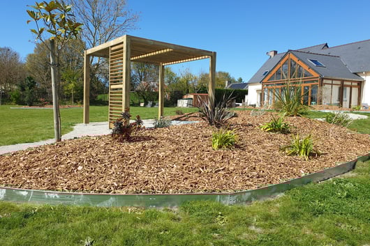 Massif paillé avec jeunes plantations devant une maison bois et pergola en fond