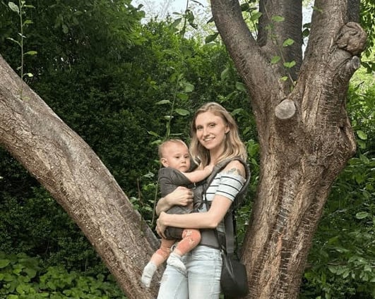 Mother holding baby in a carrier by a tree, bonding in nature.