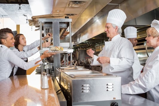 A chef hands a finished dish to a waitress in the kitchen of a restaurant.