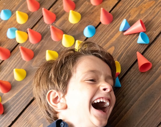 Laughing boy laying on the brown ground surrounded by blue, red and yellow silicone cones