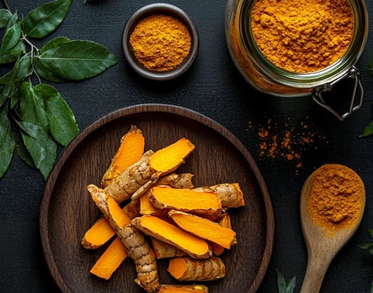 Mesa rústica vista de cima, um prato com lascas de cúrcuma e alguns potes com cúrcuma em pó
