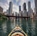 A kayak glides across a calm lake surrounded by lush green forests under a bright blue sky with scattered white clouds. The perspective from the bow of the kayak gives a sense of exploration and adventure.