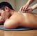 A person is lying face down with smooth black stones placed along their spine. Another person's hands are arranging the stones. Orchid flowers are present, adding a serene and natural touch to the scene. The setting suggests a spa or massage environment focused on relaxation.