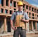 A person wearing a high-visibility safety vest and a beanie stands outdoors, operating a theodolite mounted on a tripod. The background includes the facade of a residential building with balconies.
