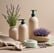 A minimalist composition featuring eco-friendly cleaning products arranged on a textured beige fabric. There's a bottle with a pump dispenser, a small packet, and two paper bags with lavender and mint green labels. The background is softly lit, creating a calm atmosphere.