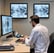 Two people in lab coats collaborate in an office setting. One is seated and pointing at a computer screen displaying medical images, while the other stands nearby holding a tablet. Large windows reveal a view of greenery outside, creating a bright and professional environment.