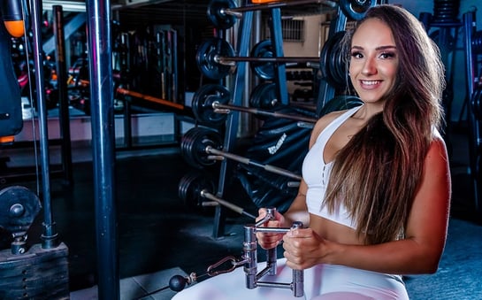 atleta de cabelos longos vestindo conjunto fitness branco e sorrindo no aparelho da academia