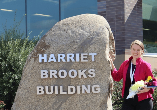 Ellen poses with flowers next to large stone sign that says "Harriet Brooks Building"