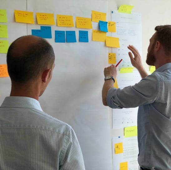 two men standing in front of a white board with sticky notes on it