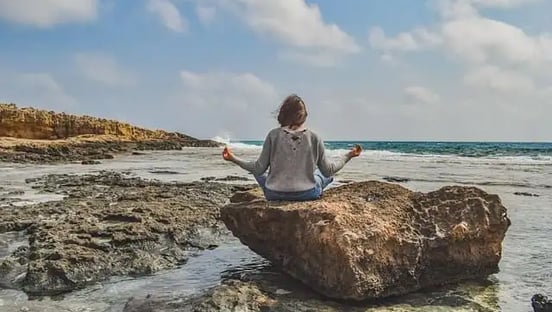 Femme assise sur un rocher face à la mer, mains écartées, doigts joints en posture de méditation