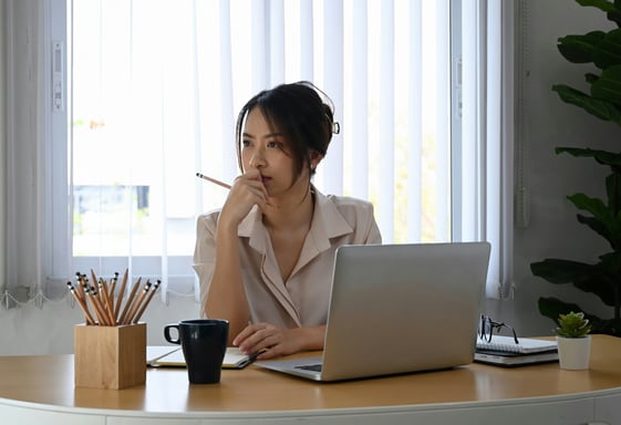 A thought young business woman sitting at a desk with an open laptop, coffee cup, and pencil holder.