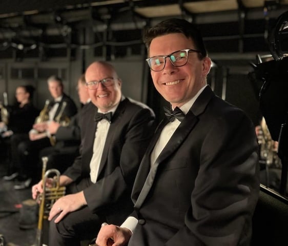 Two trumpet players wearing tuxedos, smiling in an orchestra pit.
