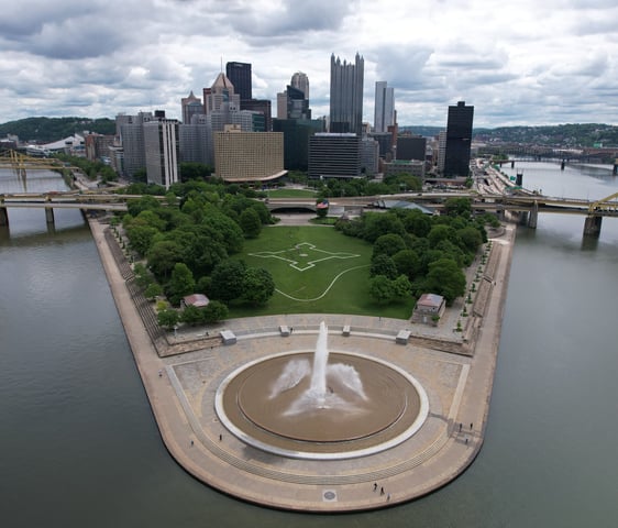 An aerial view of the Pitts Point Fountain in Pittsburgh, captured by a drone. The fountain sprays w