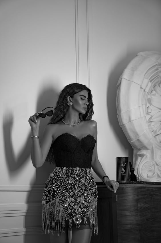 A glamorous brunette woman wearing jewelry in black lace top and sequins skirt holding black sunglasses looking to the right